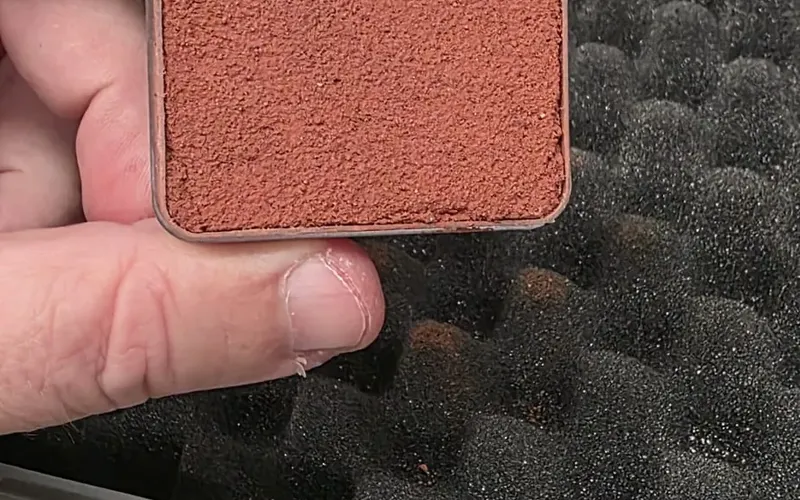 A hand holding a small brick-red sample square taken from a foam-lined sample case.