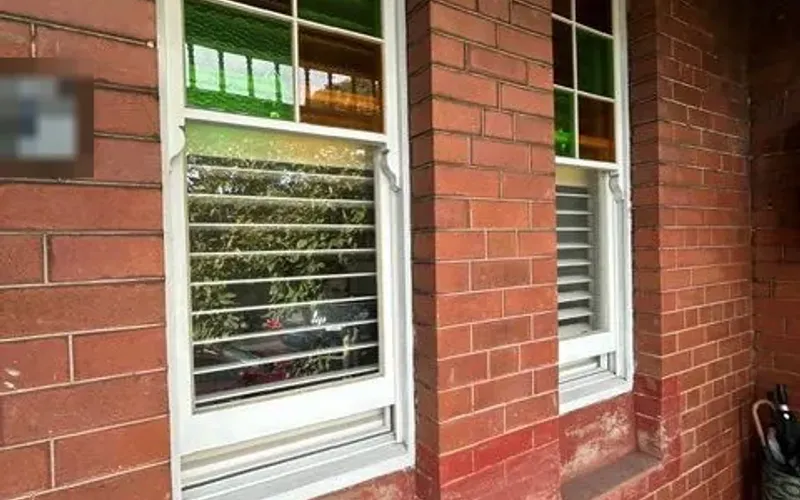 The two front windows before the work, with worn, red-painted brick sills and stained brickwork below. The edge of the house name plaque is obscured.