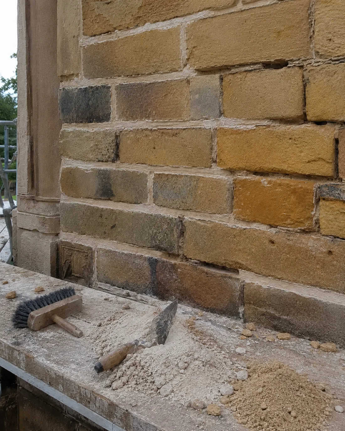 Lime mortar, trowel and brush on a plank beside a weathered sandstone wall mid-repointing